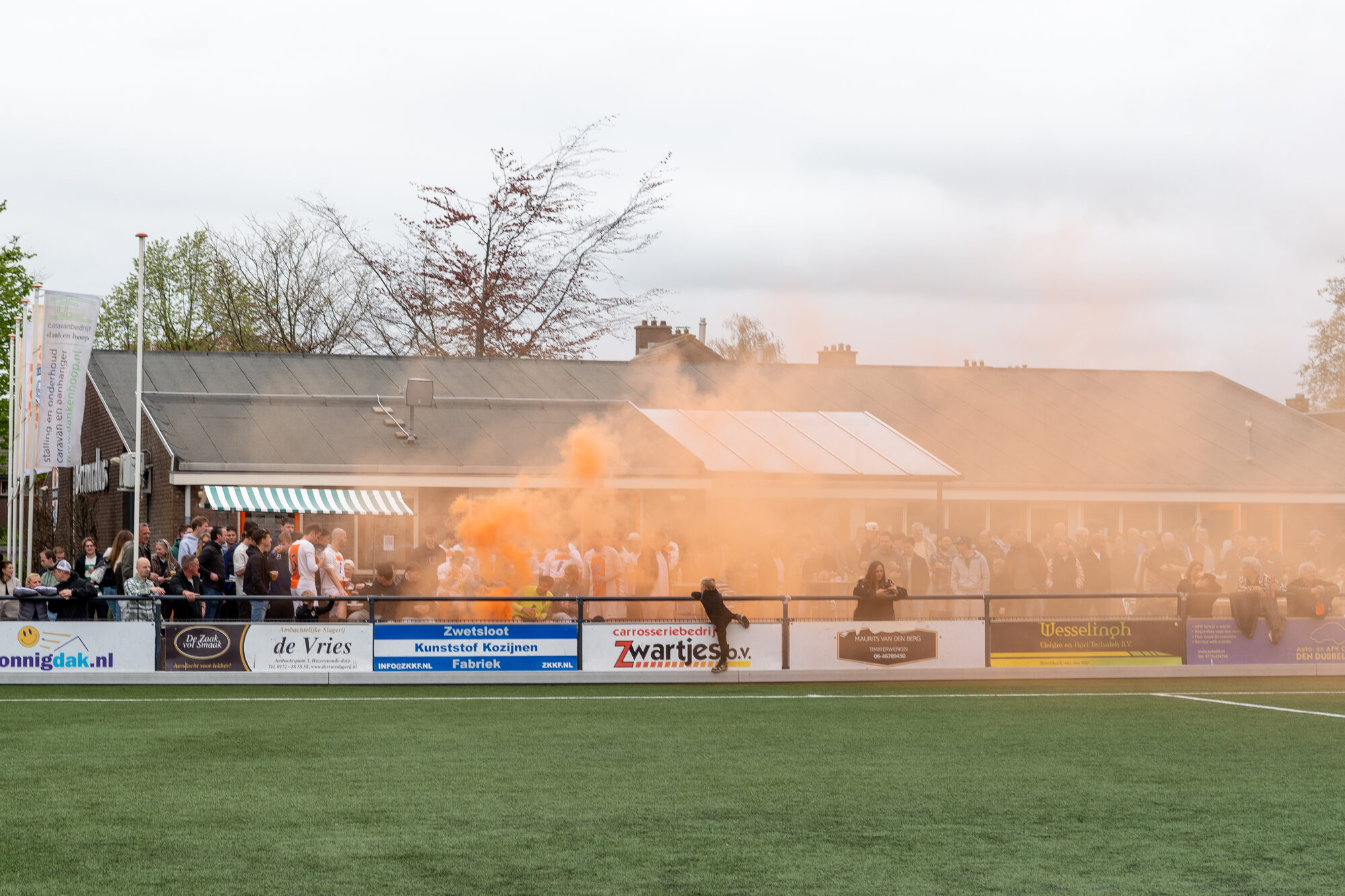 Sfeer bij het clubhuis met oranje rook langs de sponsorborden